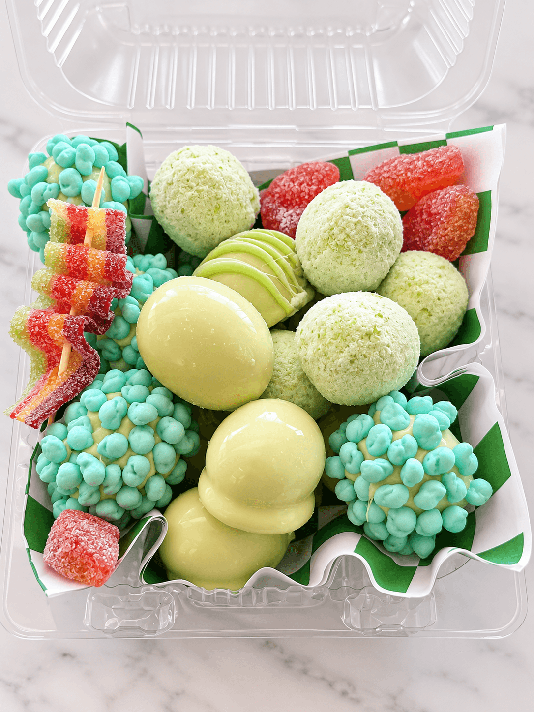Clear plastic container filled with smooth green chocolates, textured balls, teal candies, and red gummies.